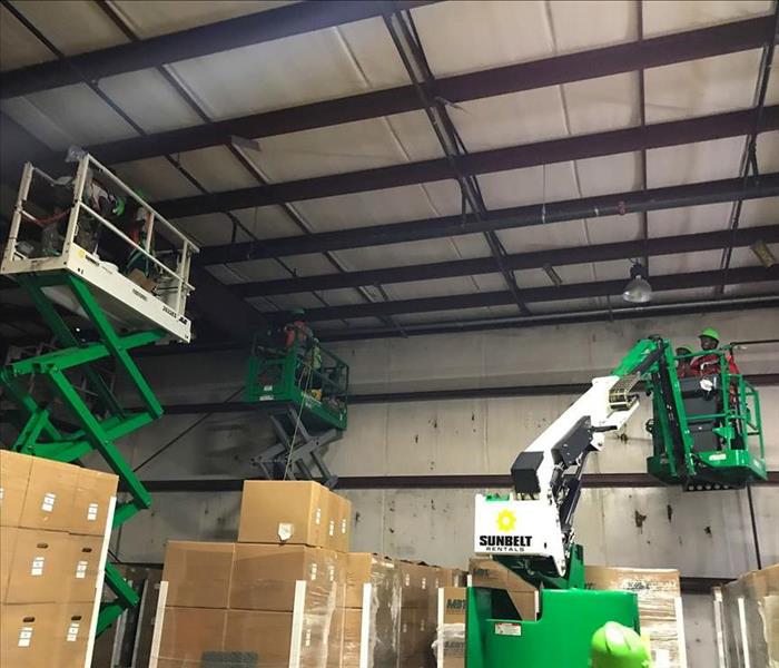 SERVPRO techs in hydraulic lifts cleaning smoke damage from the ceiling of a warehouse